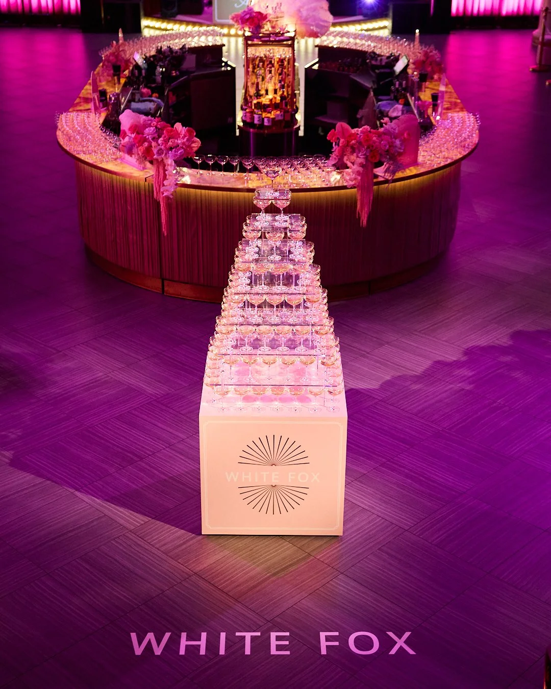 A pyramid of champagne glasses on a table with the logo 'White Fox', at a decorated event space with pink and purple lighting, floral arrangements, and a circular bar in the background.