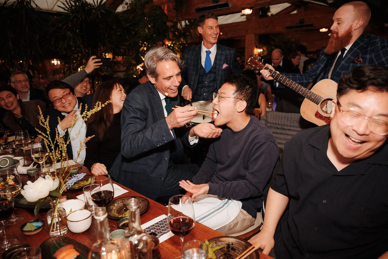 People enjoying a celebration with food, drinks, and live music in a cozy, warmly lit indoor setting.
