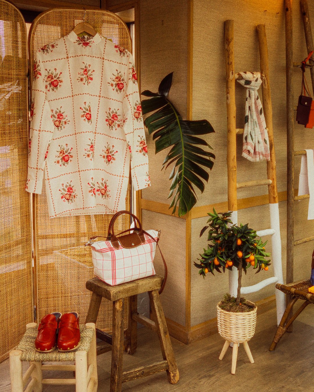 A display of fashion and decor items including a white dress with red floral pattern, a plaid bag on a wooden stool, red shoes on a small woven stool, a large green leaf, a small potted orange tree, a wooden ladder with a scarf, and a woven chair wit