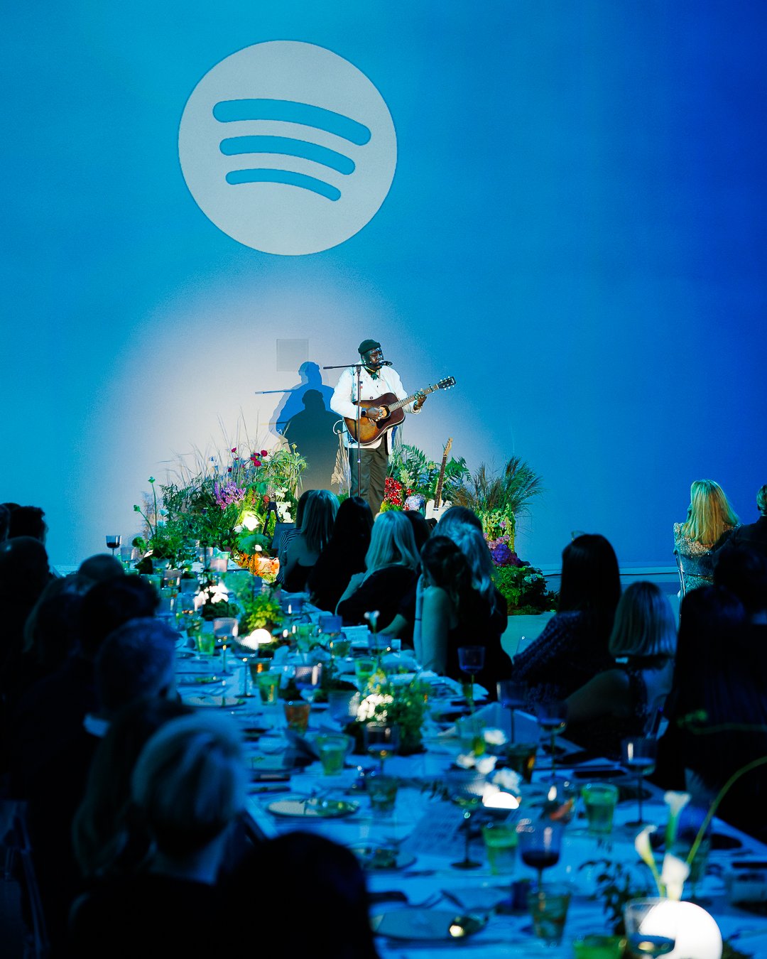 Musician performing on stage with guitar in front of an audience at an indoor event, with a large Spotify logo projected on the blue wall behind him, decorated with flowers, tables, and chairs filled with guests.