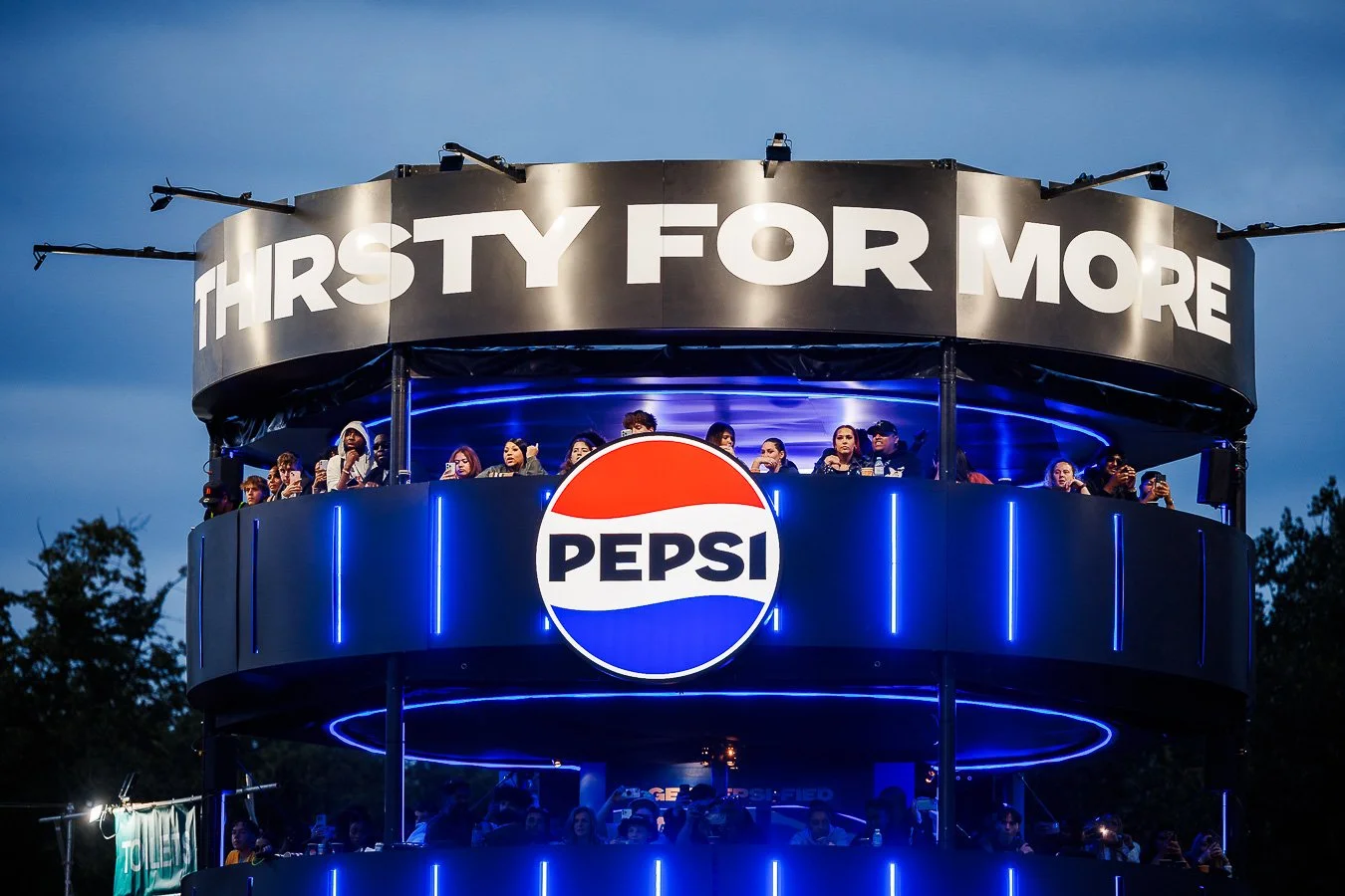 An outdoor amusement park ride with two levels, a Pepsi logo at the center, and a large sign reading 'Thirsty for More' at the top, with people sitting and enjoying the ride during early evening.