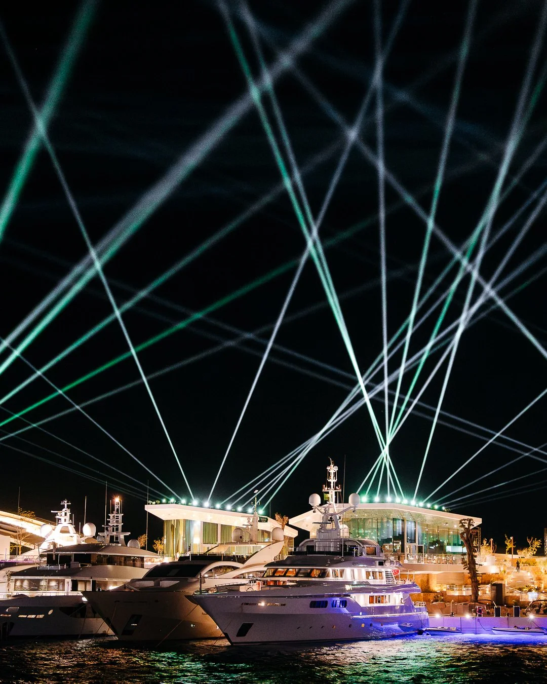 Night scene at a marina with yachts docked beside a modern building illuminated with green lights. Spotlights project green beams into the night sky, creating a crisscross pattern.