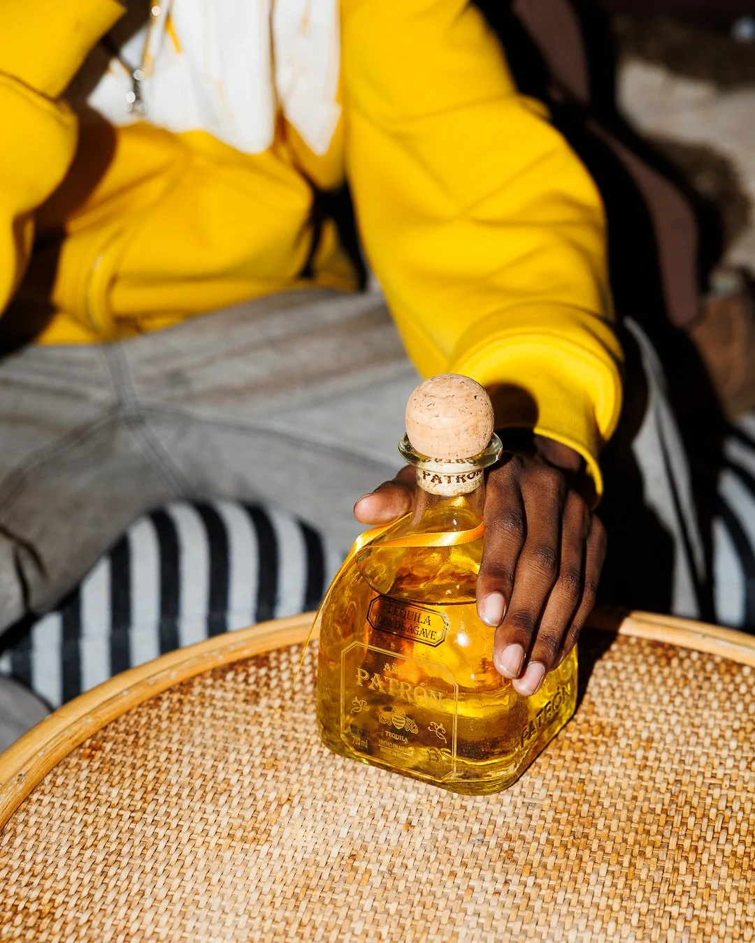 Person wearing a yellow jacket holding a bottle of Patrón tequila on a woven table.