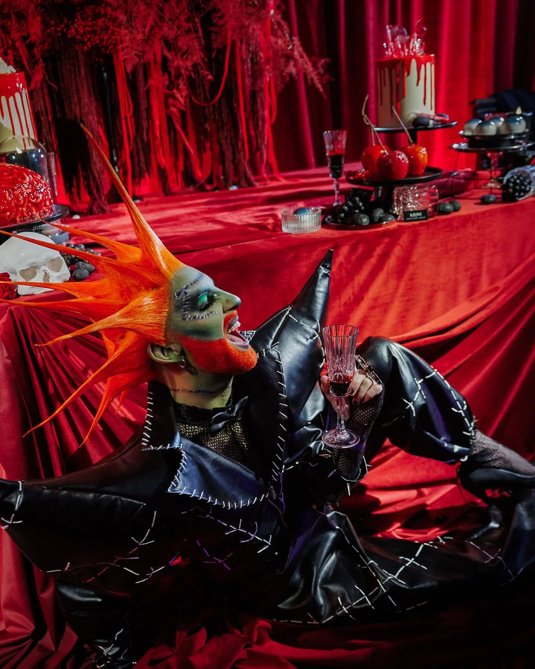 A person with vibrant orange spiked hair, dramatic makeup, and a black leather outfit sitting on a red cloth-covered surface, holding a glass of red wine, with a colorful, gothic-styled party setup in the background.