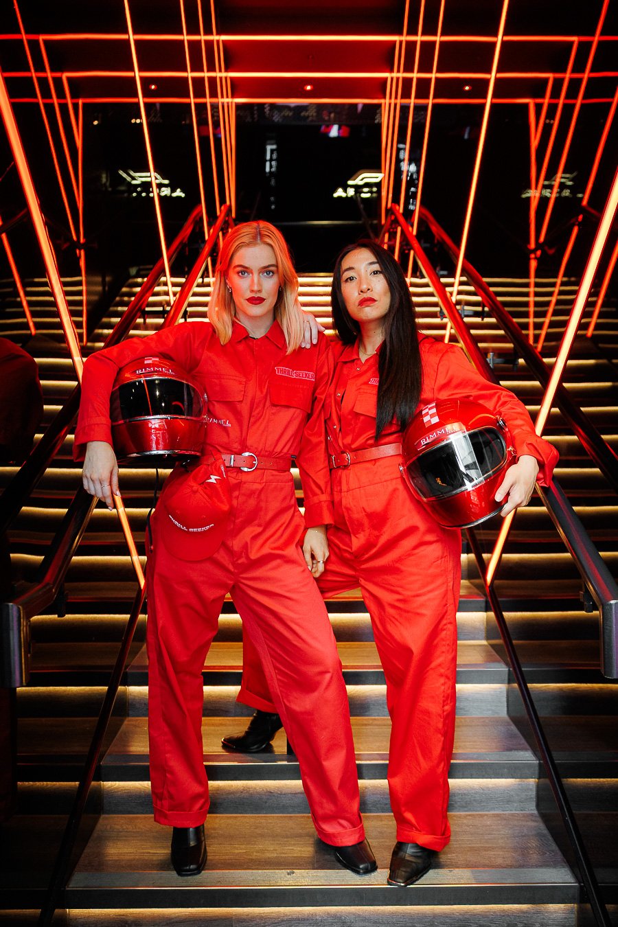 Two women in red racing suits holding helmets, standing on a staircase with neon lights overhead.