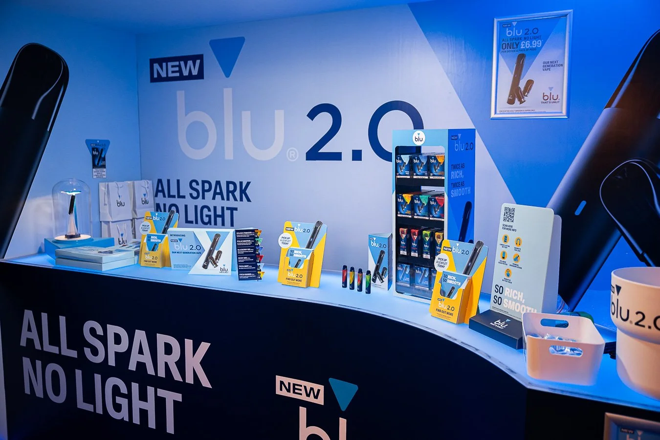 Display booth showcasing the Blu 2.0 vaping device, with promotional signs and product samples on the table. The backdrop features the Blu 2.0 logo and slogan "All Spark No Light".