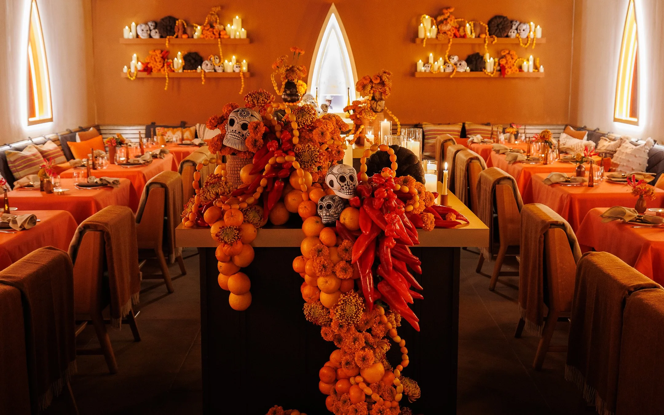 Decorated dining room with orange tablecloths, candles, and intricate floral arrangements. The centerpiece features skull-shaped decorations, orange and red chili peppers, and bright orange fruits, accented by orange flowers. Warm lighting and candle