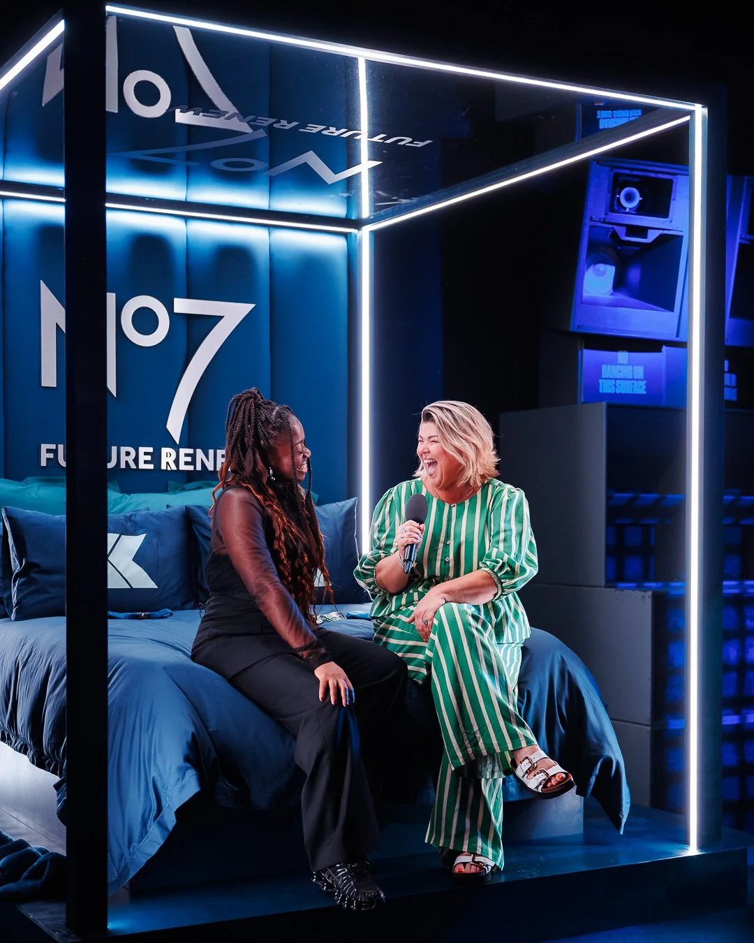 Two women sitting on a bed with blue bedding, laughing and talking. One holds a microphone. The background features illuminated signage with the number 107 and the words "FUTURE REN". The setting has modern, neon lighting.