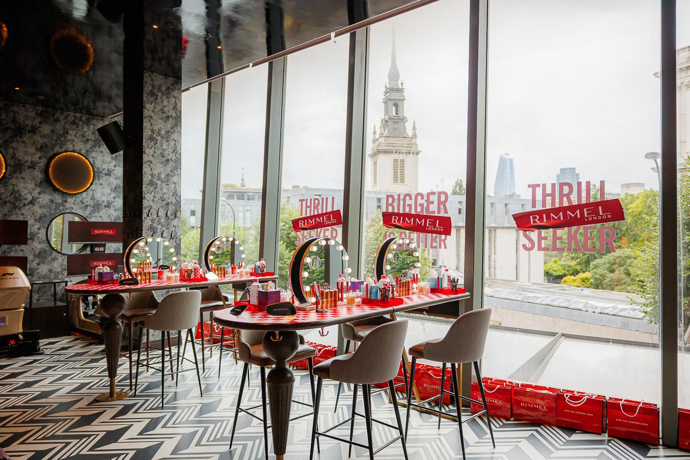 Beauty station with makeup mirrors, products, and chairs inside a modern salon with large windows showing city skyline and promotional posters.