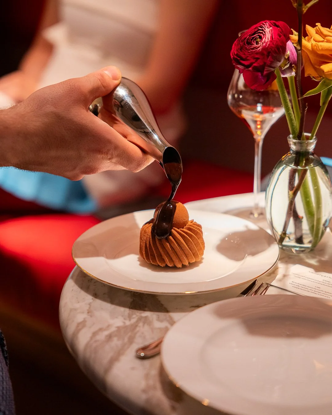 A person is pouring chocolate sauce over a dessert on a white plate at a fancy dining table, with a vase of flowers and empty plates nearby.