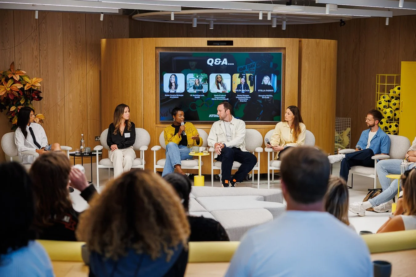 Panel discussion with six diverse speakers on stage, seated in white chairs, engaging with each other in a well-lit room with an audience in front. Large screen behind them shows a Q&A session.
