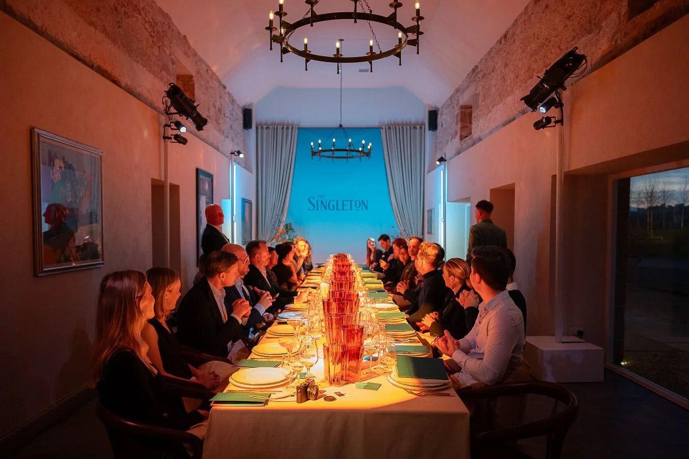 A large family or group of people gathered around a long dinner table in a rustic-style dining room with exposed brick walls and a vaulted ceiling. There are 20 adults seated, with a man standing at the head of the table. The table is set with plates
