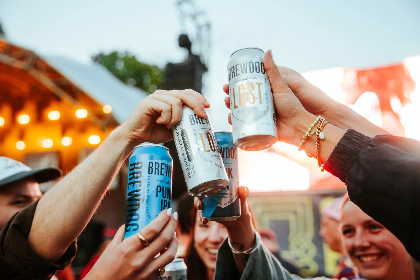 Group of people raising cans of Brewdog beer at an outdoor event, with a blurred background of lights and a sunset.