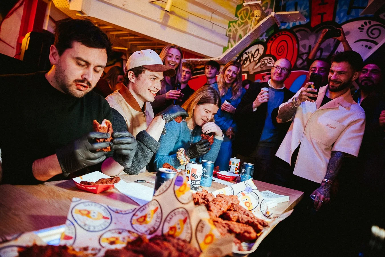 People gathered around a table enjoying fried chicken at a party with colorful graffiti artwork in the background.