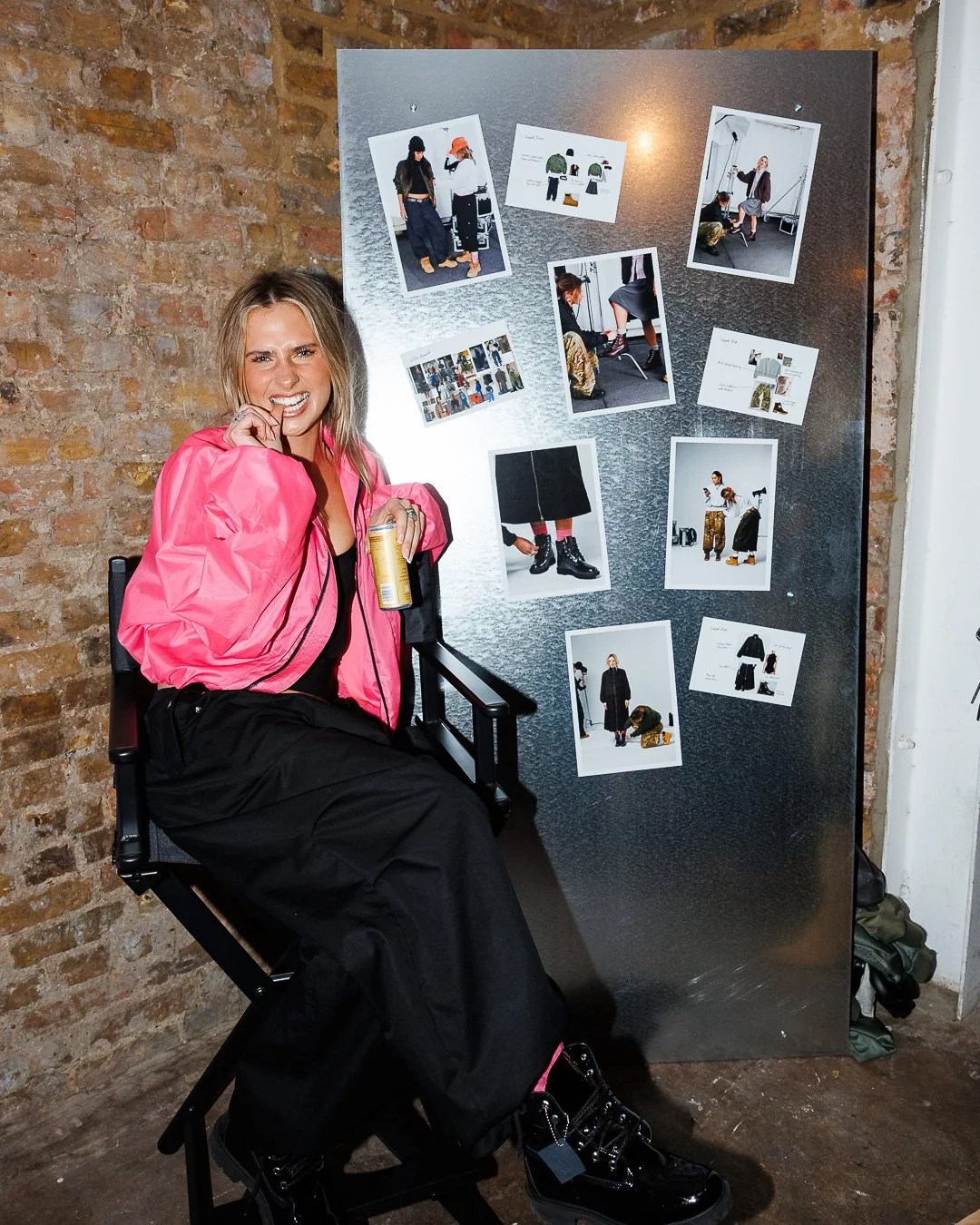 A woman sitting in a director's chair smiling, holding a can, wearing a pink windbreaker and black pants, next to a display board with photos and sketches of fashion designs against a brick wall background.