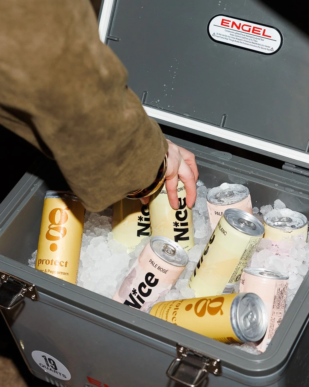Person opening a gray cooler filled with ice and assorted canned beverages, including LaCroix sparkling water and hard seltzers.