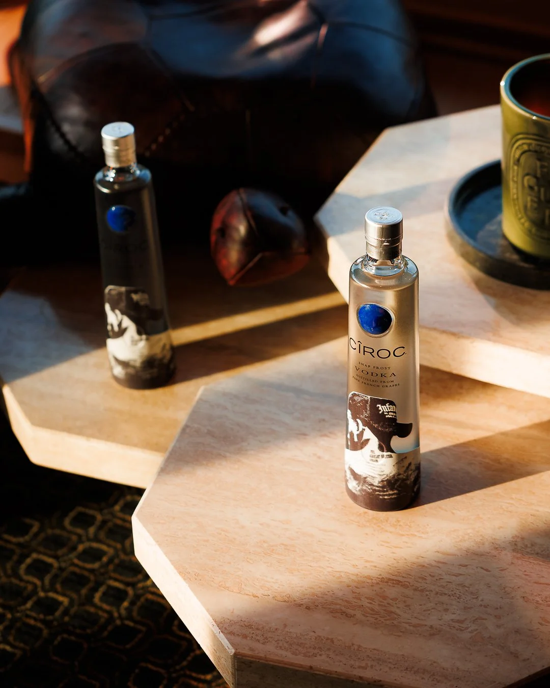 A bottle of Ciroc vodka on a wooden table with sunlight casting shadows, a black bag, a green mug on a tray, and a leather basketball in the background.