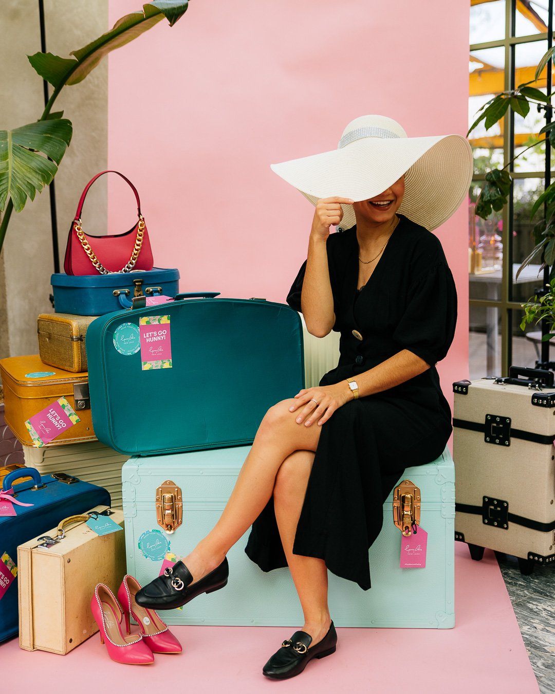 Woman sitting on a large blue suitcase, wearing a black dress and a wide-brimmed white hat, surrounded by colorful suitcases and handbags in a setting with plants and pink background.