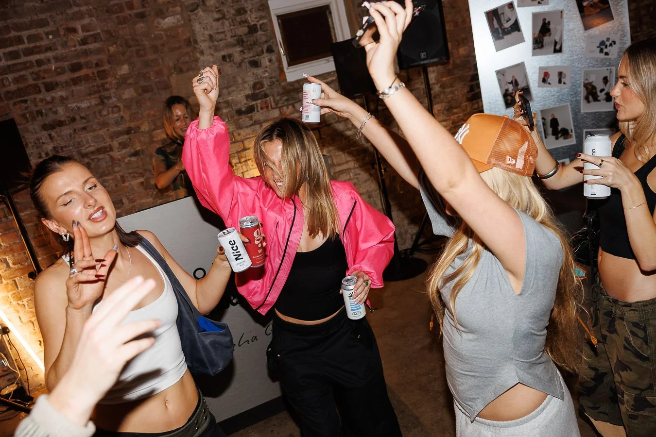 Group of women dancing and having fun at a party, with drinks in hand, in a brick-walled venue.