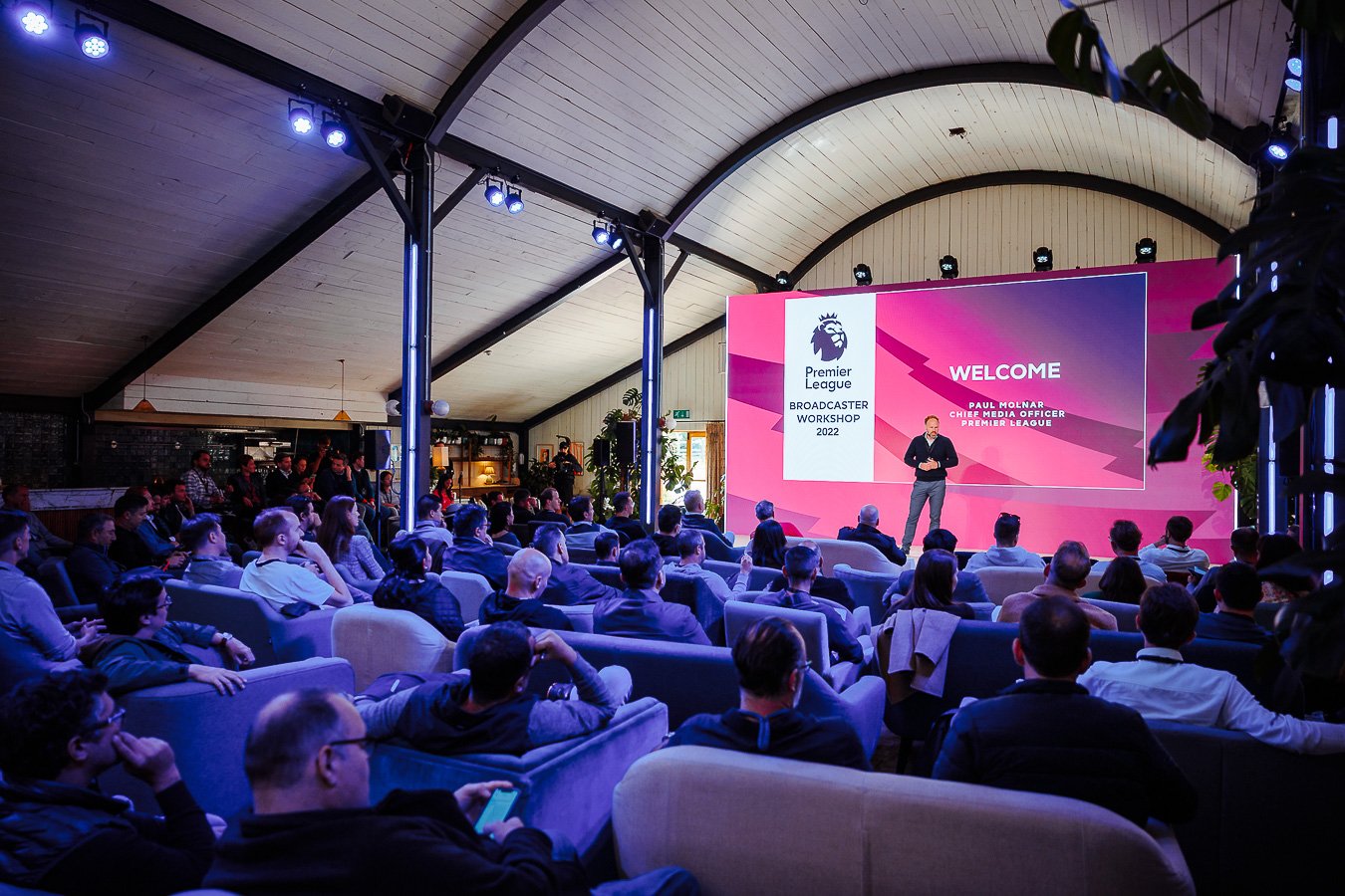 A speaker presenting at a conference or workshop in a large room with a pink and purple screen behind him displaying the Premier League logo and the text welcome and details of the event. Attendees are seated and listening.