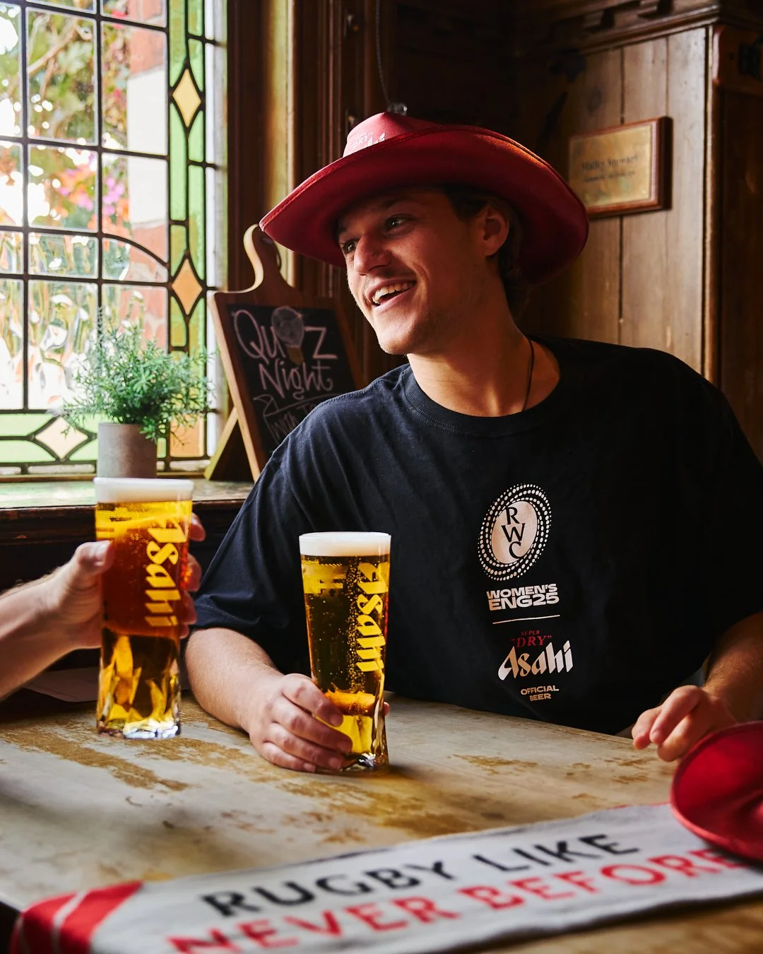A young man in a red hat and black T-shirt holding a glass of beer, smiling and engaging with others at a pub or brewery.