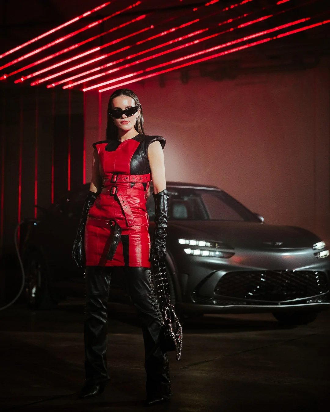 A woman in a red and black leather outfit with long dark gloves and sunglasses, standing in front of a sleek gray car with red neon lights overhead.