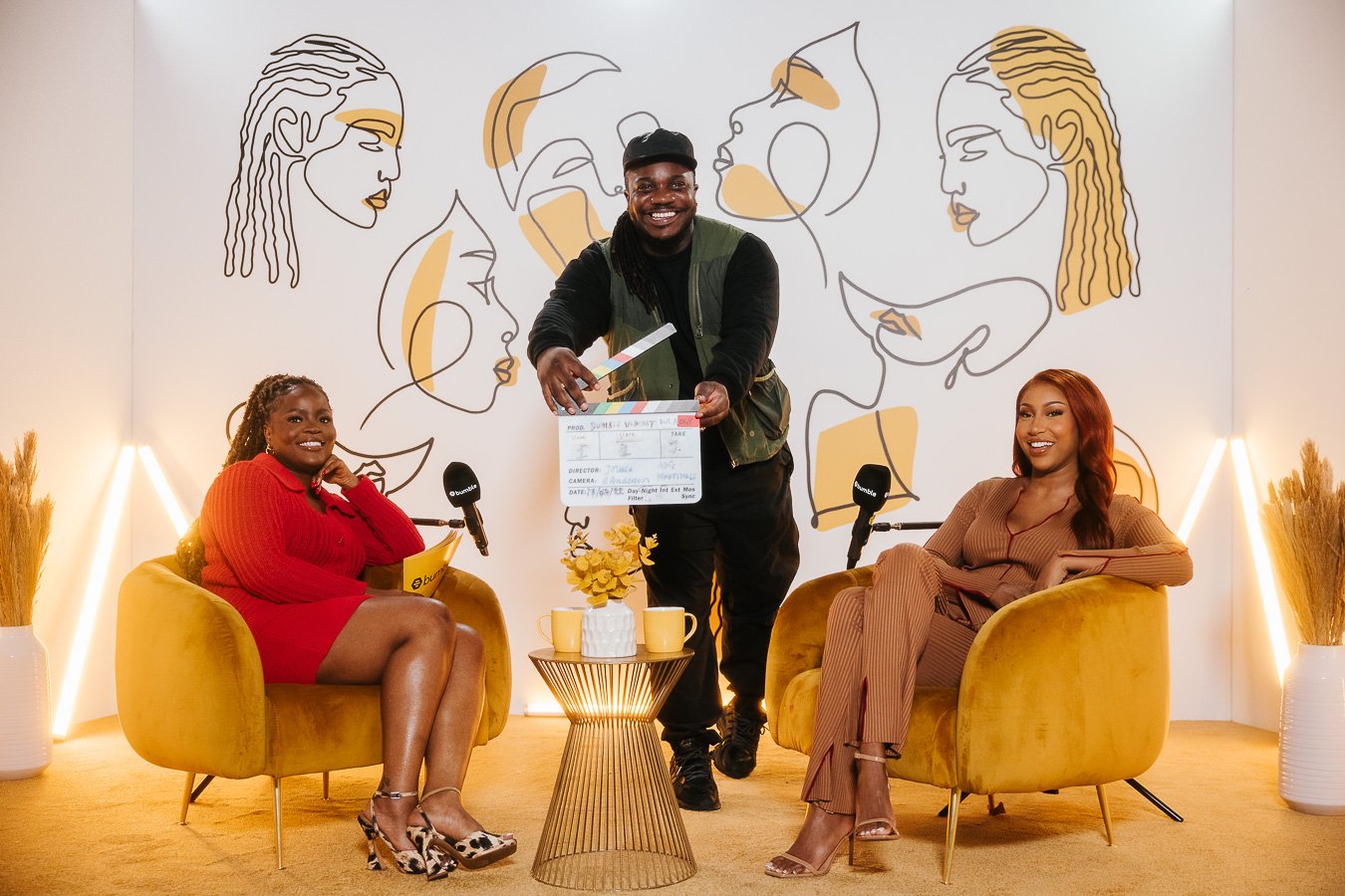 People participating in a podcast recording, with a man holding a clapboard and two women sitting in yellow chairs, in a room decorated with abstract line art of women on the wall.