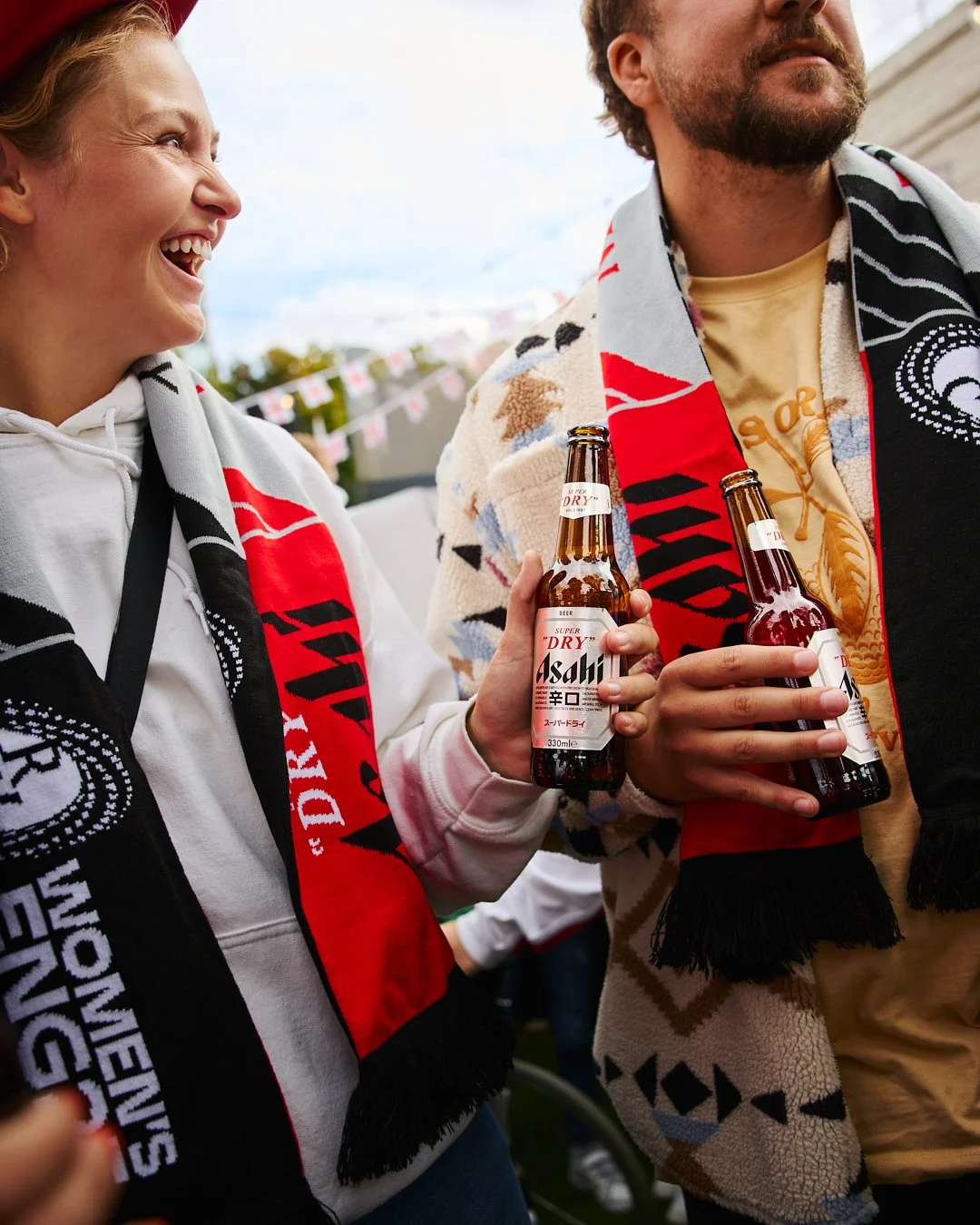 Two people holding bottles of Asahi beer and smiling, wearing fan scarves at an outdoor event.