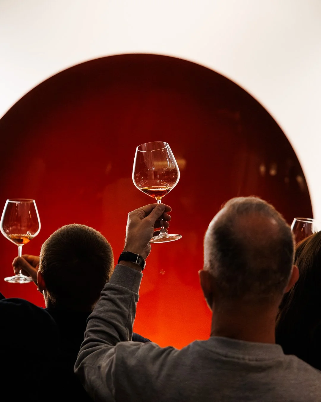 People at a wine tasting event, raising glasses of wine against a red background with a circular opening.