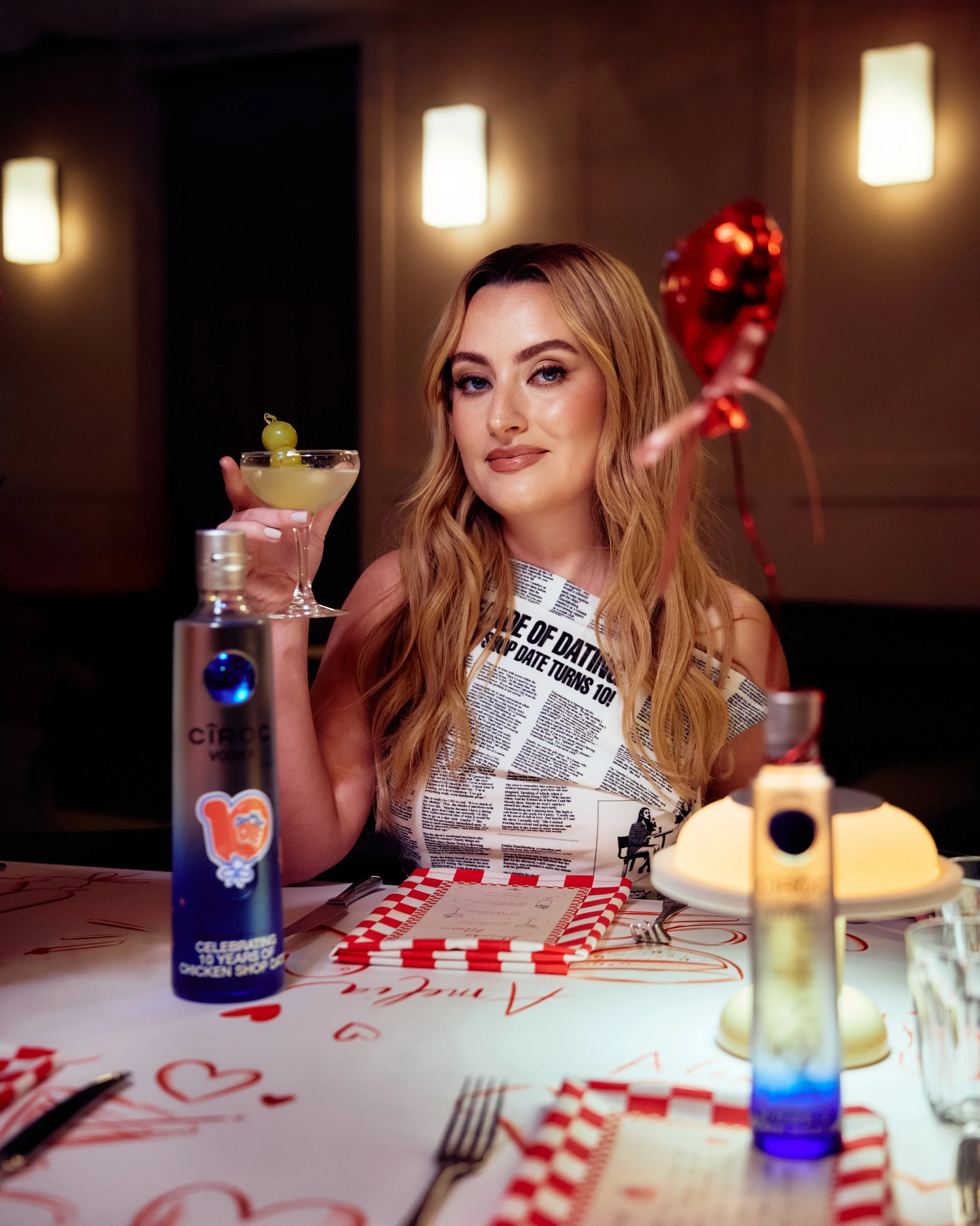 A woman with long red hair and makeup sitting at a decorated table in a restaurant, holding a cocktail glass with a lime garnish, surrounded by birthday decorations and drinks.
