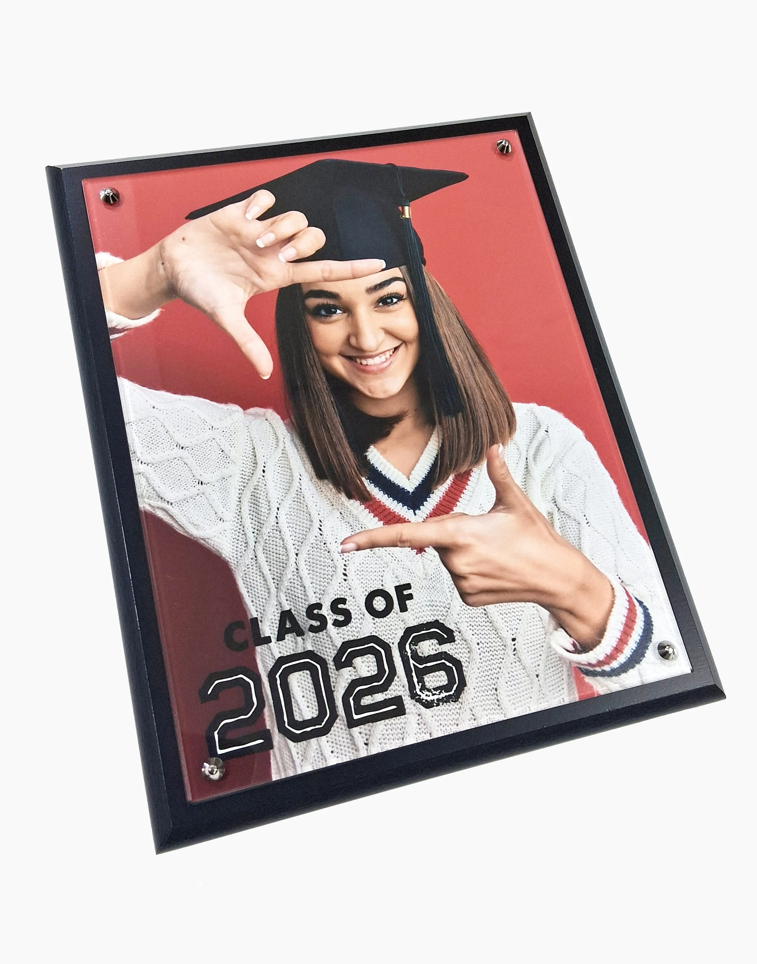 Graduation photo featuring a smiling young woman in a white sweater with red and blue striped trim, wearing a black academic cap with a tassel, making a thumbs-up gesture with her right hand, and pointing at the text on a sign that reads 'Class of 20