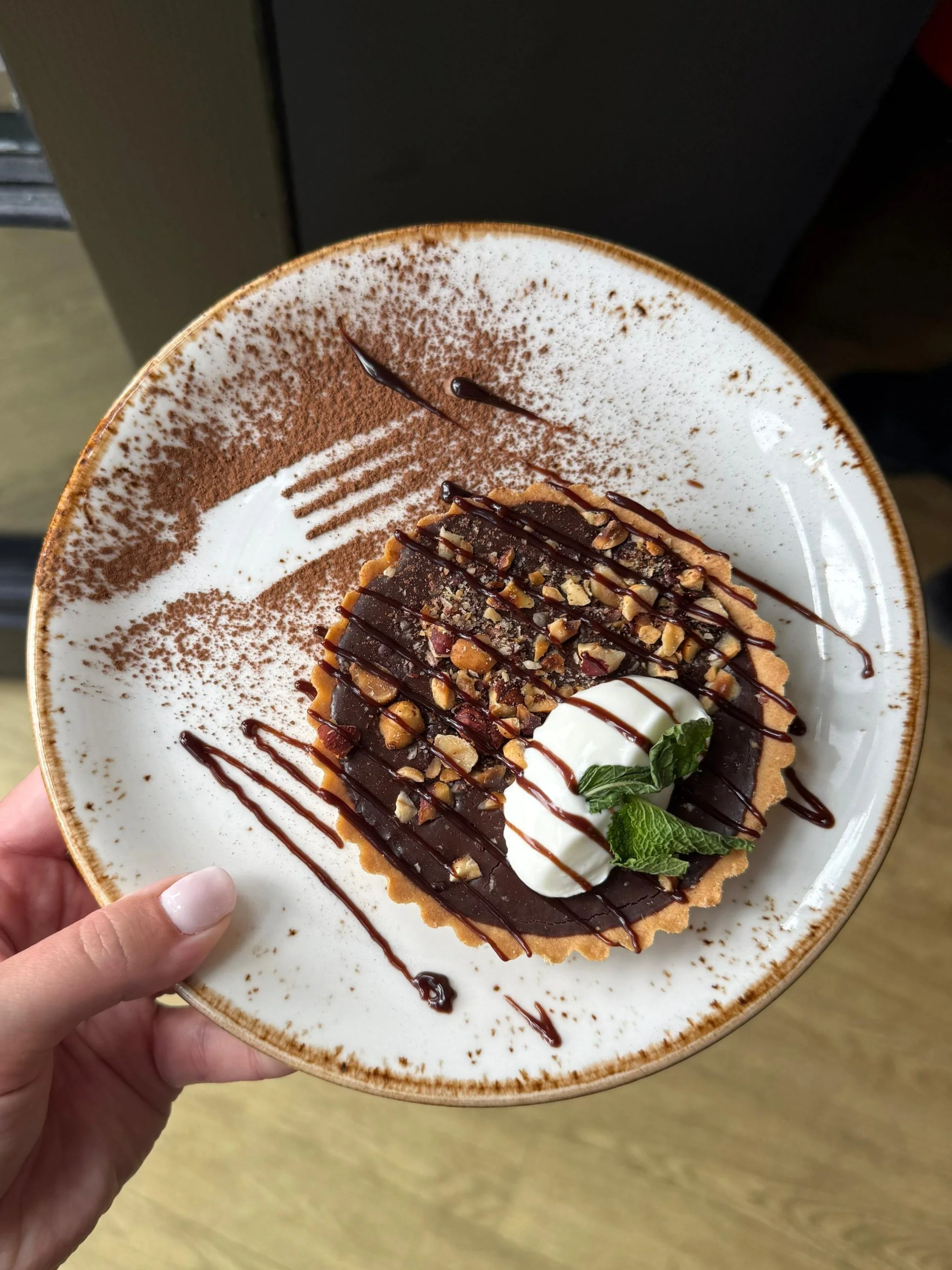 A Chocolate tart topped with hazelnuts, crème fraiche, a mint leaf, and chocolate drizzle, on a white speckled plate.