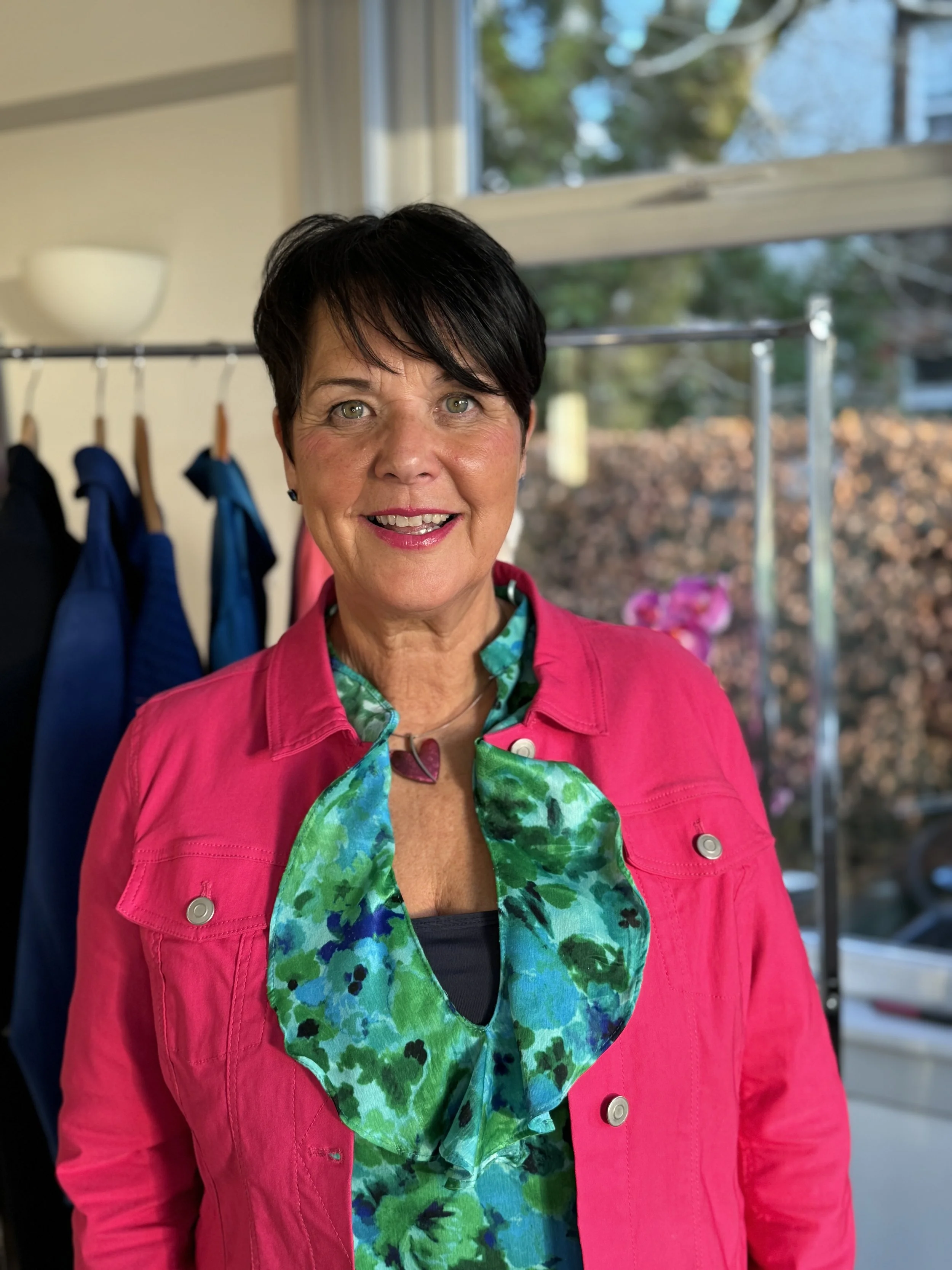 A woman with short dark hair—Shirley—wearing a bright pink jacket over a green and blue floral top, smiling indoors near a window with a blurred outdoor background. This is imagery for Colours by Shirley.
