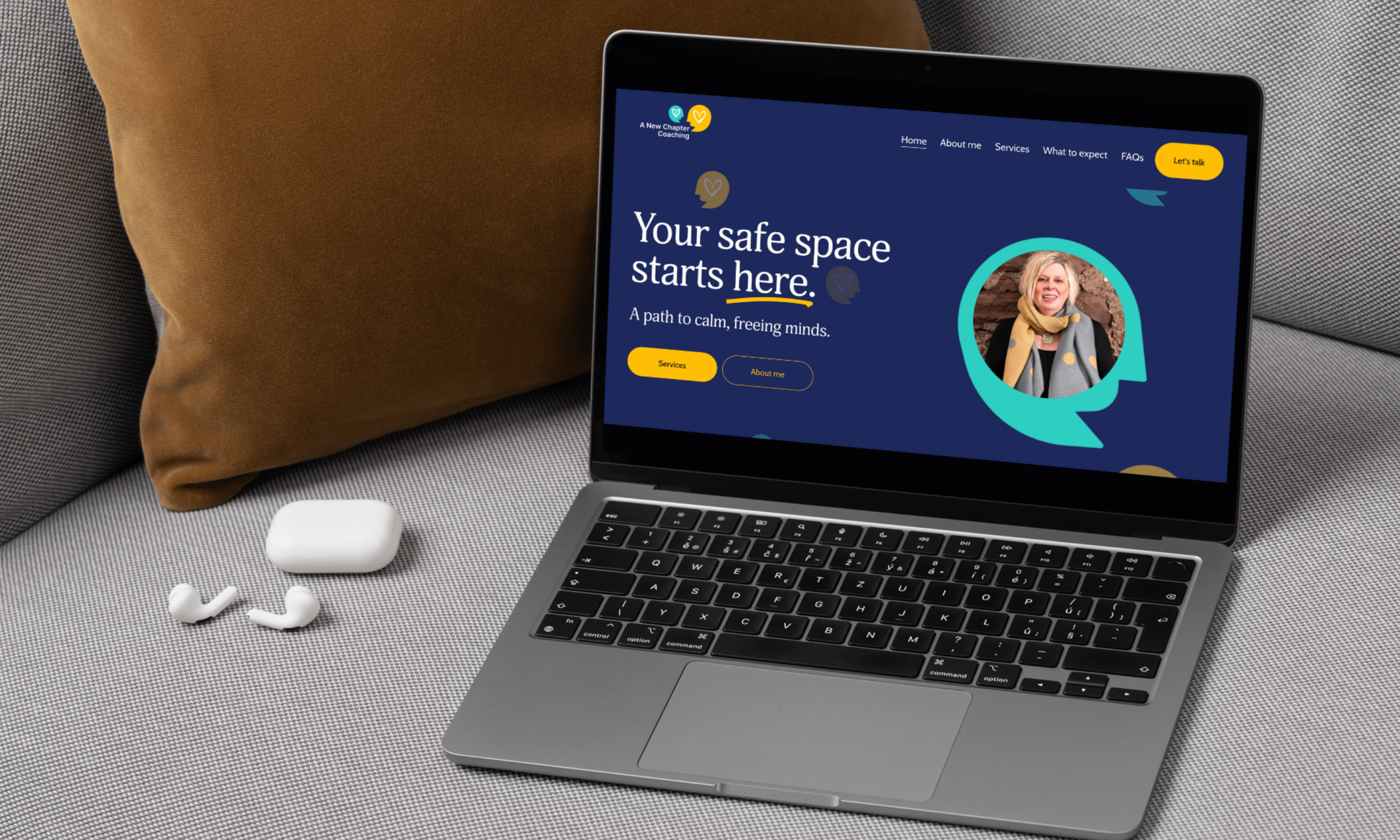 A laptop on a gray textured surface displaying a website titled 'Your safe space starts here' with a photo of a woman in a yellow scarf. Next to the laptop are white wireless earbuds, a white case, and a brown pillow.