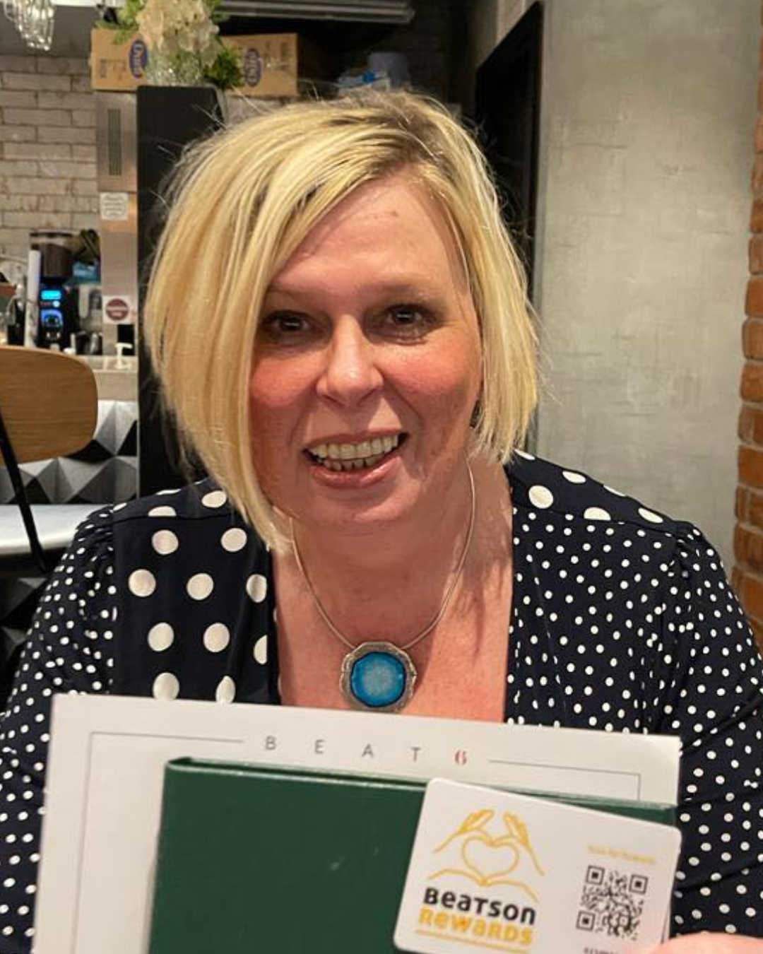 A woman with blonde hair smiling at a restaurant, wearing a black polka dot top and a blue pendant necklace, holding a menu with a 'Beatson Rewards' loyalty card.