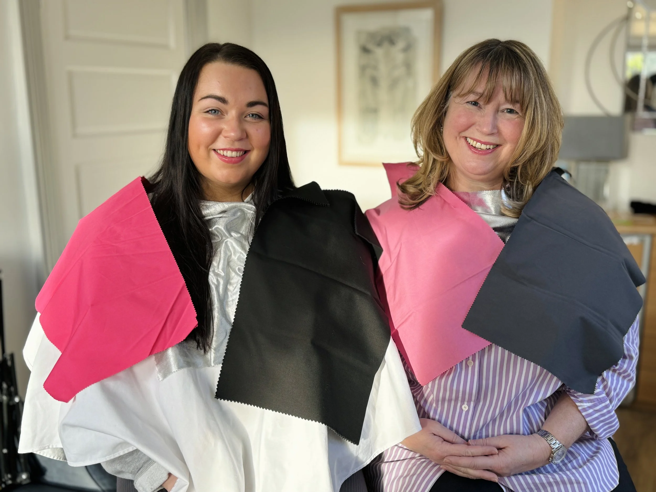 Two women with white and colourful swatches over their shoulders, smiling indoors with artwork and furniture in the background.