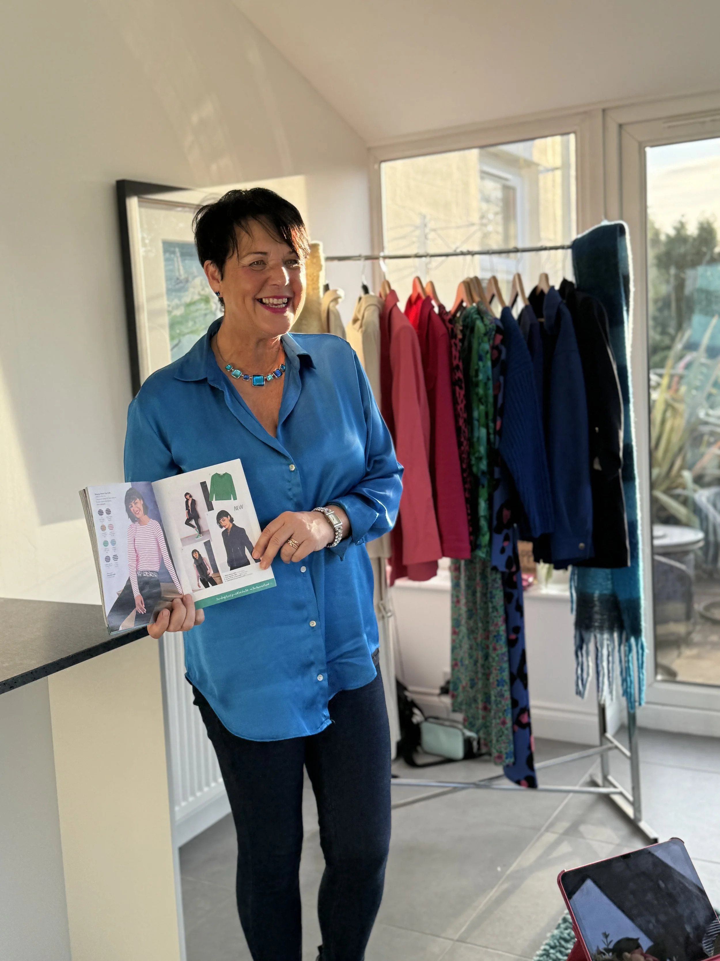 A woman with short dark hair and a bright smile, wearing a blue blouse and dark trousers, standing in front of a clothing rack with colourful clothing, holding a brochure, in a well-lit room with large windows. This is imagery for Colours by Shirley.