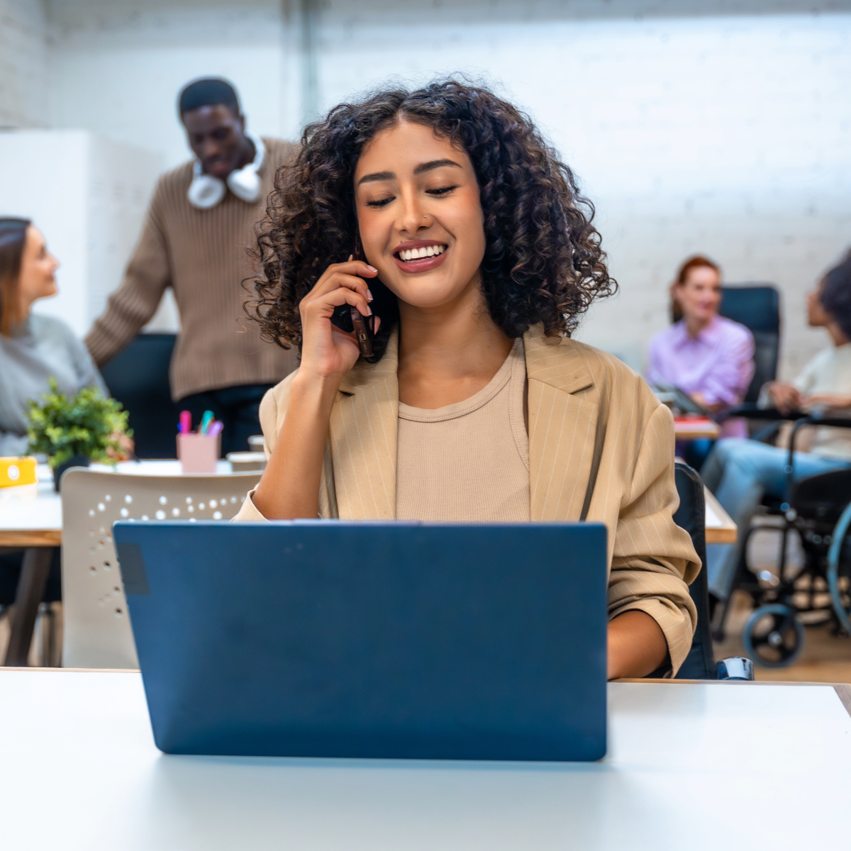 A woman is sitting at the front of the image. She is on a blue laptop and is also on the phone. She's got curly shoulder-length hair and is smiling. In the background are other workers.