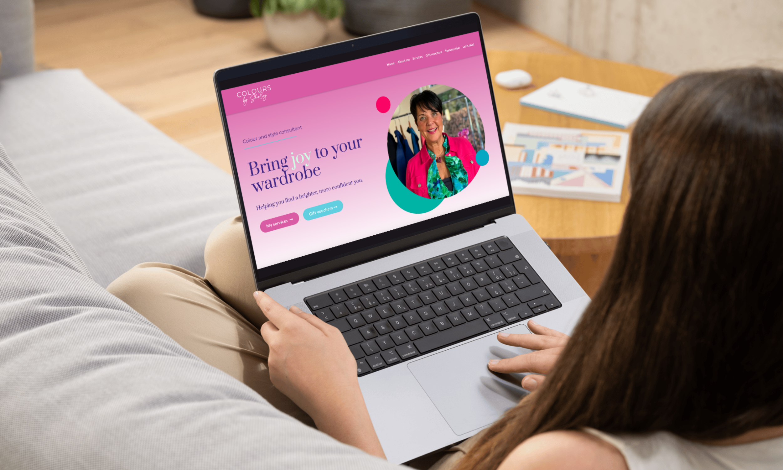 A woman sitting on a sofa using a laptop, viewing Colour by Shirley's website. The website homepage shows an image consultancy with a pink and purple theme, featuring a photo of a woman in colourful clothing.