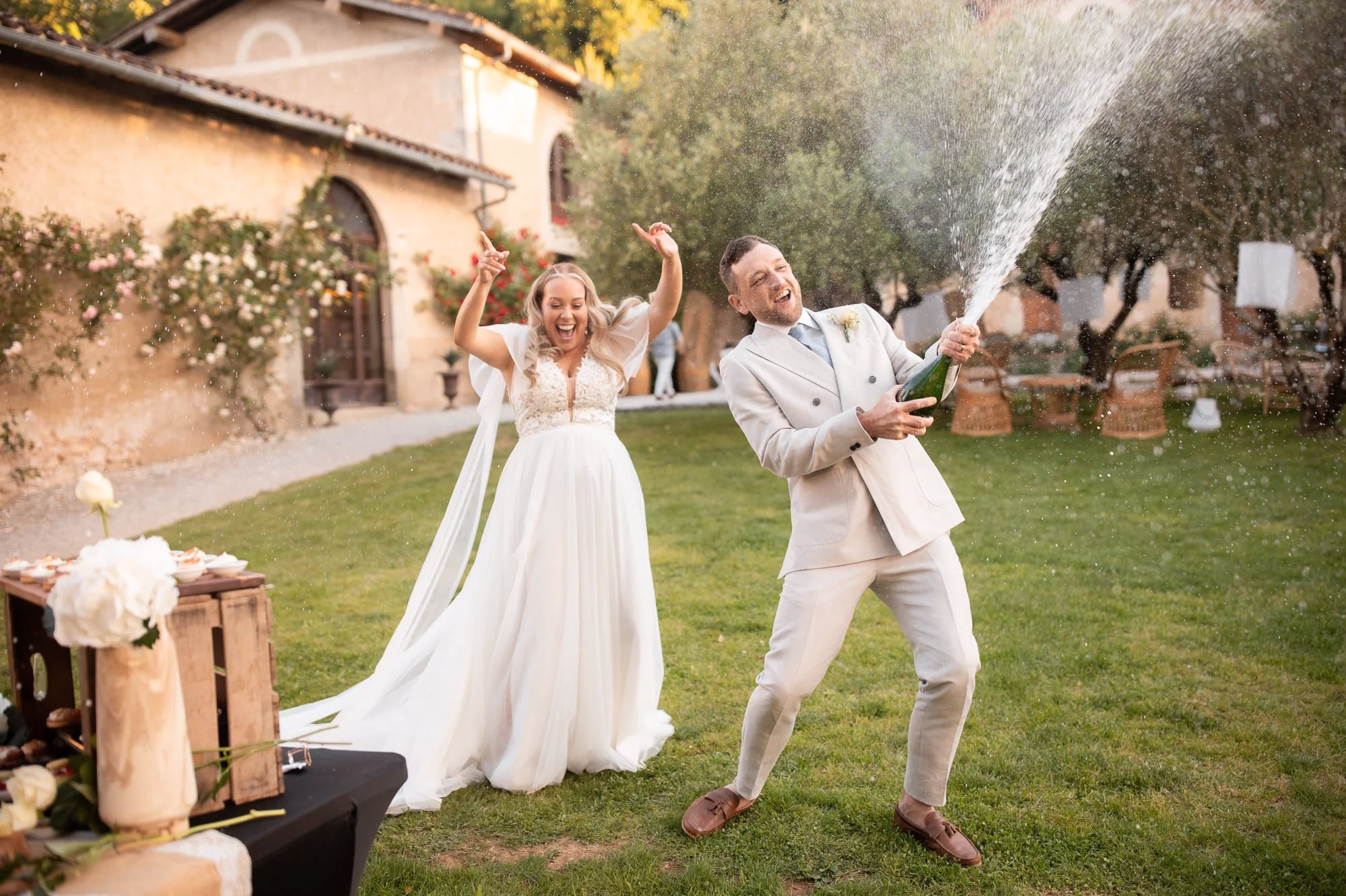 A bride and groom celebrating outdoors at a wedding reception, with the groom opening a champagne bottle while the bride dances behind him, both smiling and enjoying the moment on a lush green lawn with tables and trees in the background.