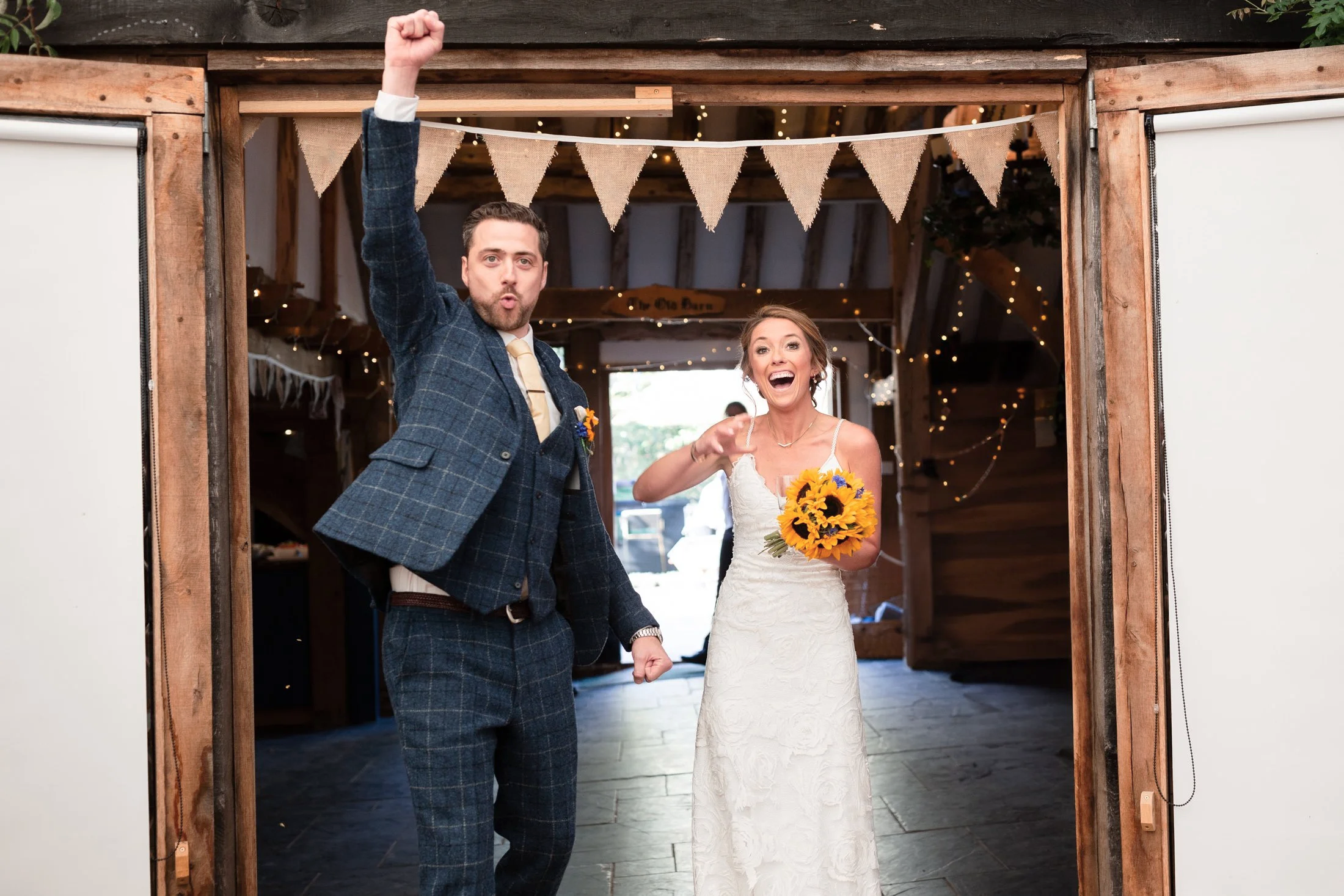 A man and woman celebrating at a wedding, standing in a rustic wooden venue decorated with bunting and string lights. The man has his arm raised and makes a fist, while the woman holds a bouquet of sunflowers and appears excited and happy.