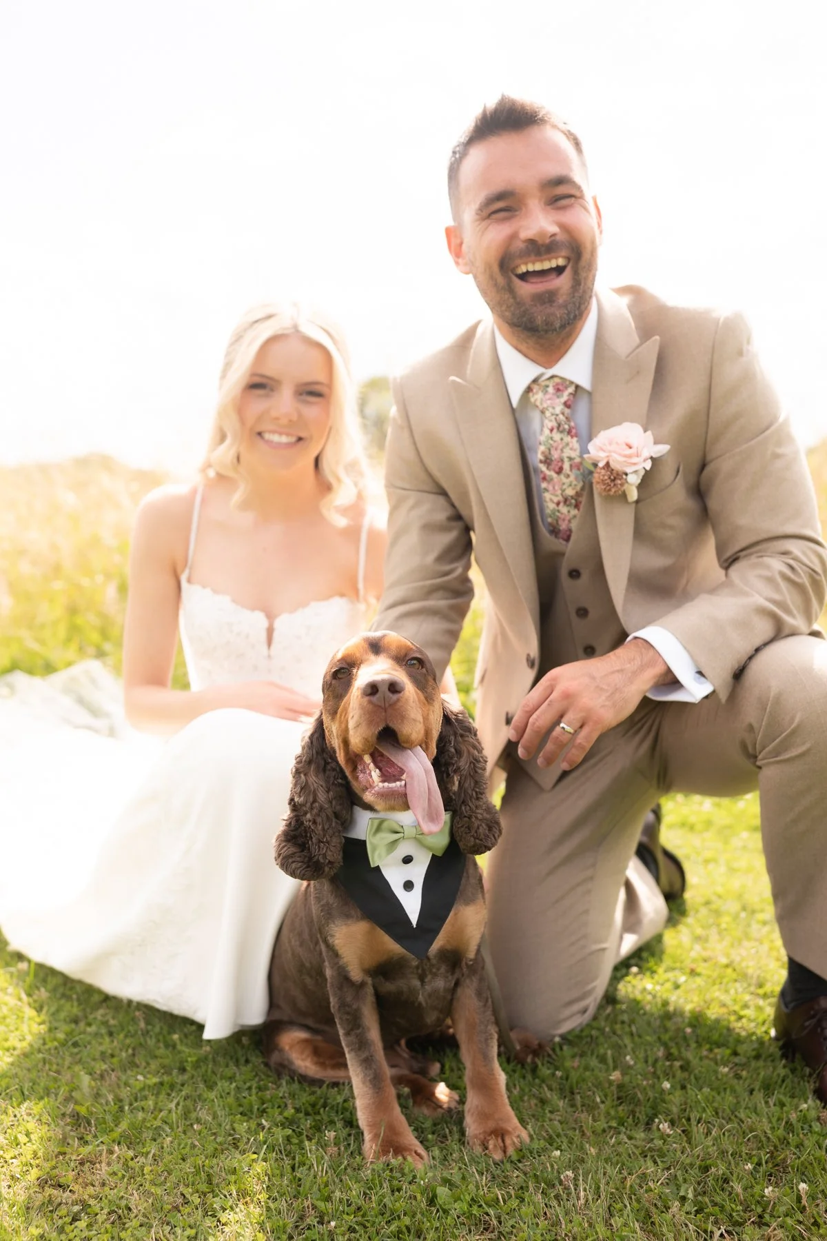 A smiling couple, a woman in a white dress and a man in a beige suit with a floral tie and boutonniere, along with a happy brown and black cocker spaniel dog wearing a tuxedo bow tie, outdoors on grass during daytime.