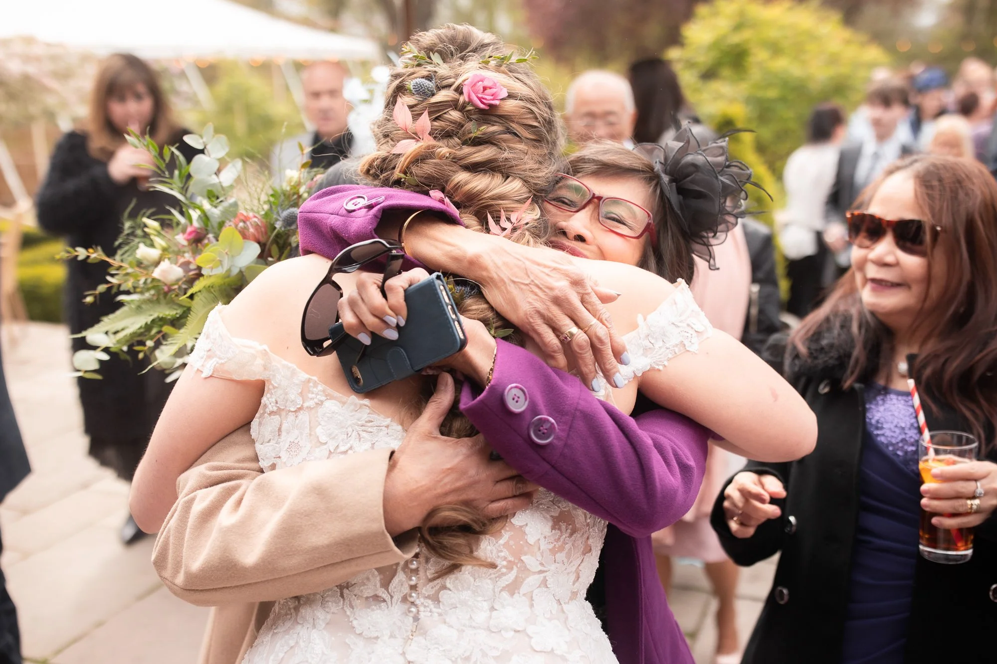 Two women embracing in a hug at an outdoor event, with other people in the background, some holding drinks, and trees with autumn foliage.