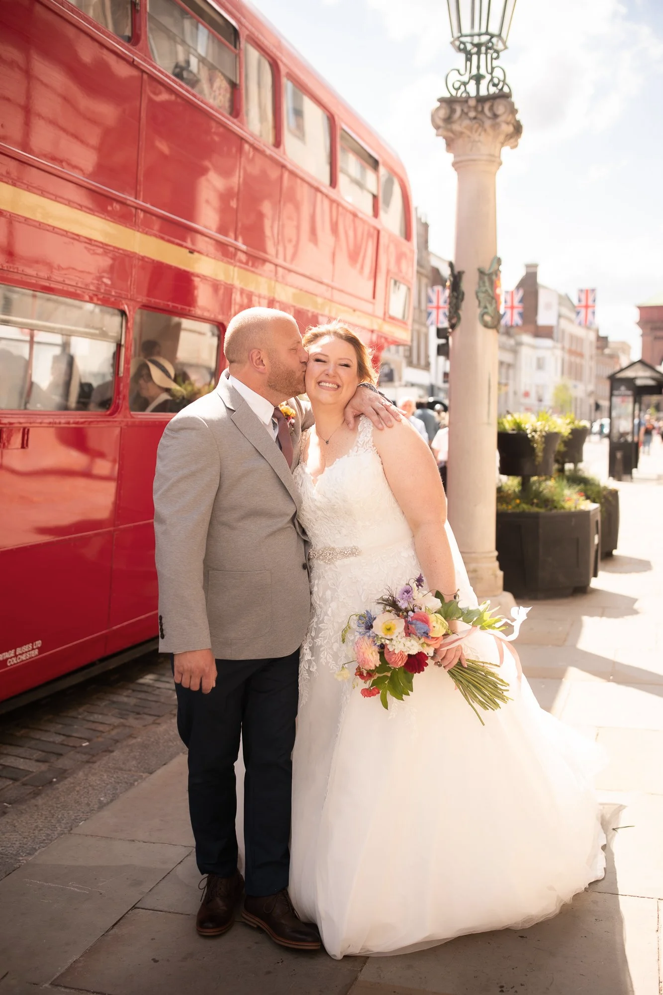 A bride and groom sharing a kiss on a sunny day in a city street. The bride is holding a colorful bouquet and wearing a white wedding gown. The groom is dressed in a gray suit. Behind them is a red double-decker bus and Union Jack flags, with histori