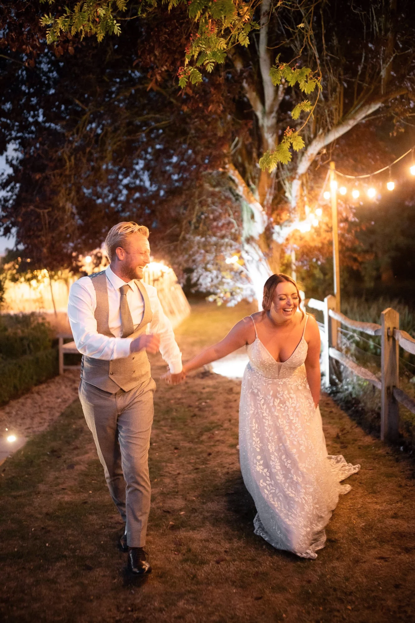 A bride and groom are walking hand in hand outdoors at night, smiling and laughing, with a large illuminated tree and string lights in the background.