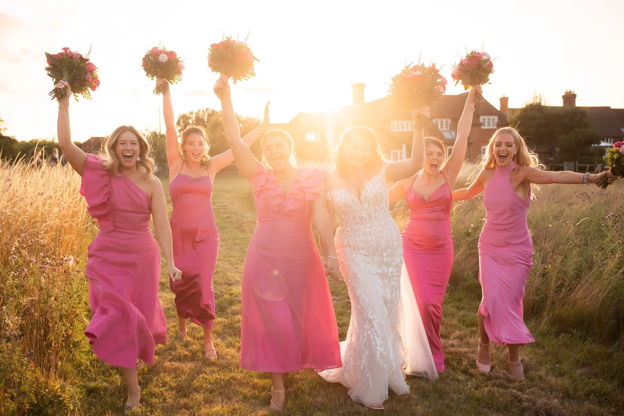 A bridal party of six women in pink dresses and a bride in a white gown celebrating outside during sunset, holding bouquets and smiling.