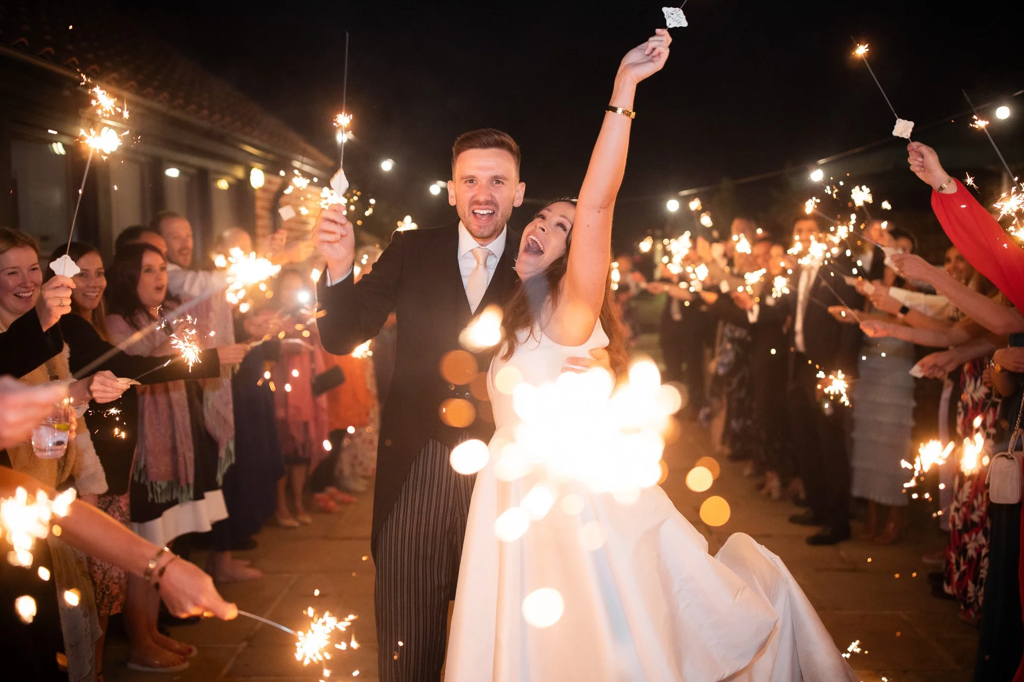 A newlywed couple dancing at their wedding reception surrounded by guests holding sparklers at night.
