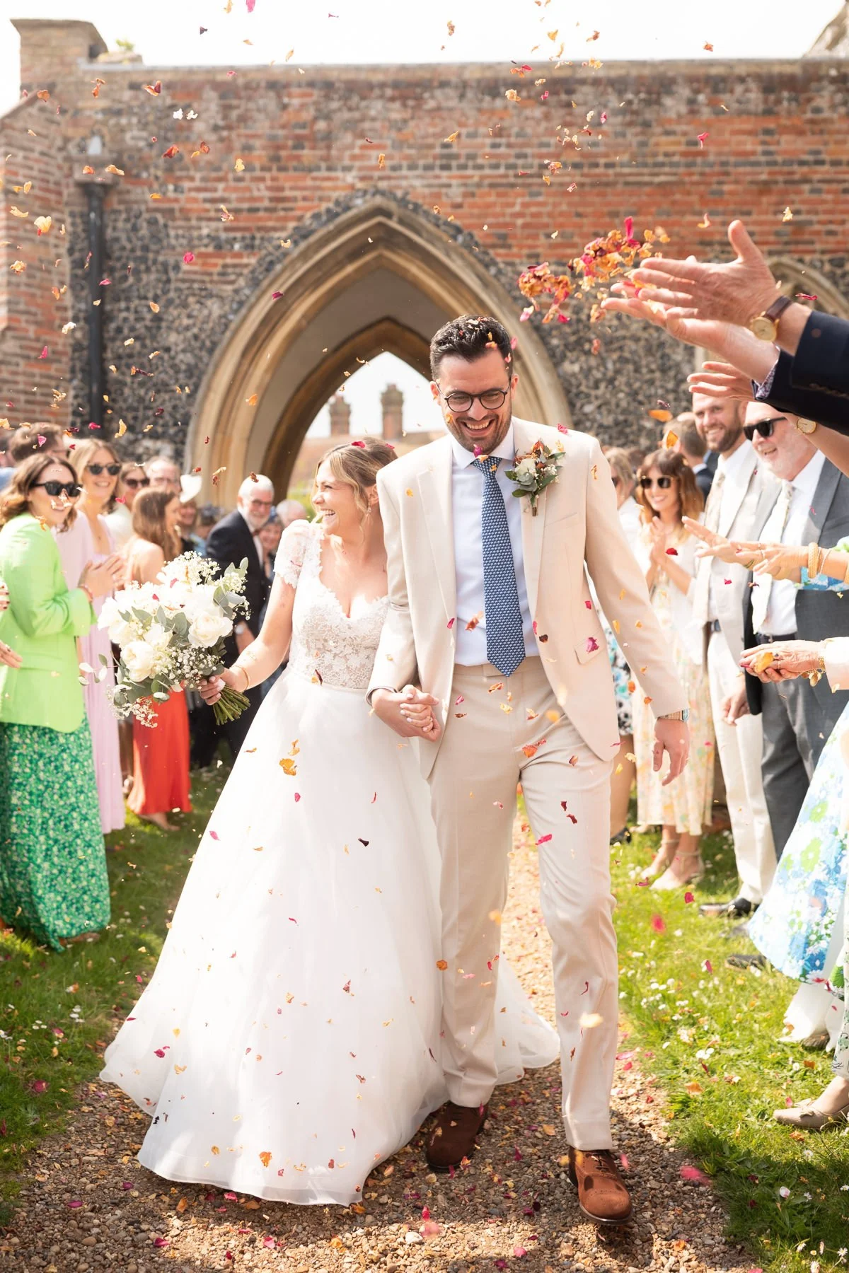 A newlywed couple walking hand in hand through a crowd of wedding guests outside a historic stone building, surrounded by falling flower petals during daytime.