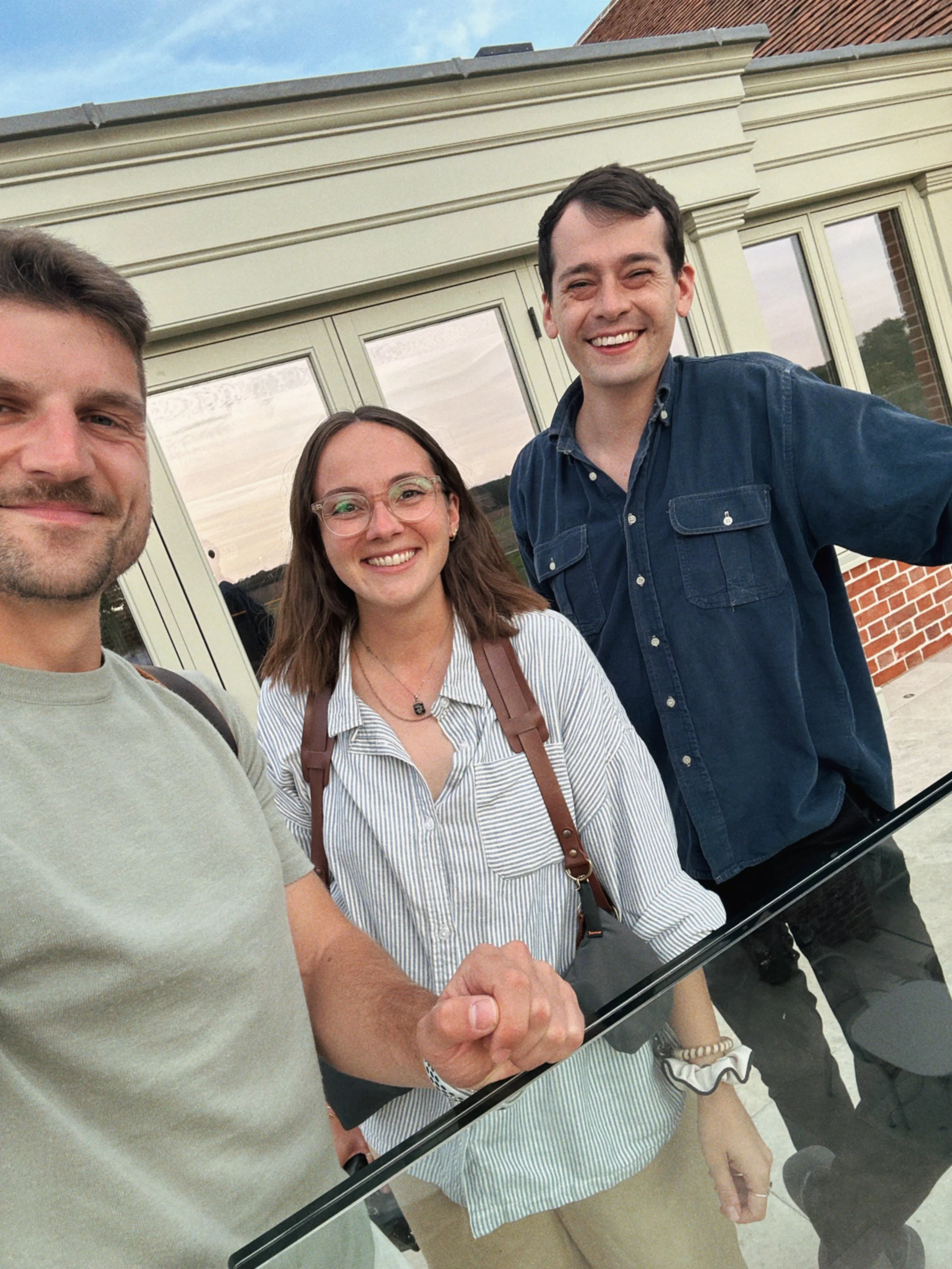 Three smiling people taking a selfie together outdoors in front of a building with large windows and brick accents.