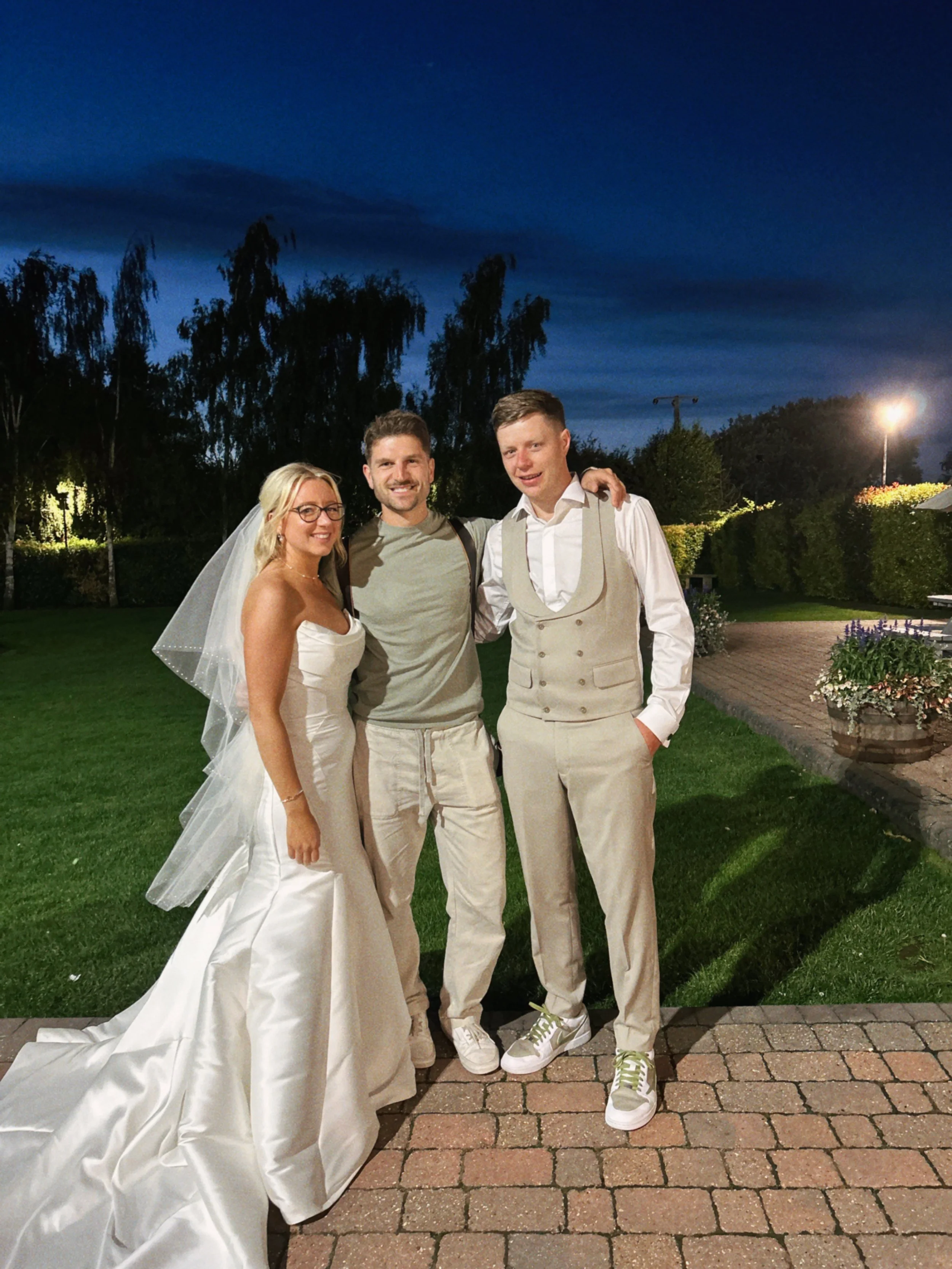 A bride and two men standing together outdoors at night, with trees and a lit area behind them, during a wedding celebration.